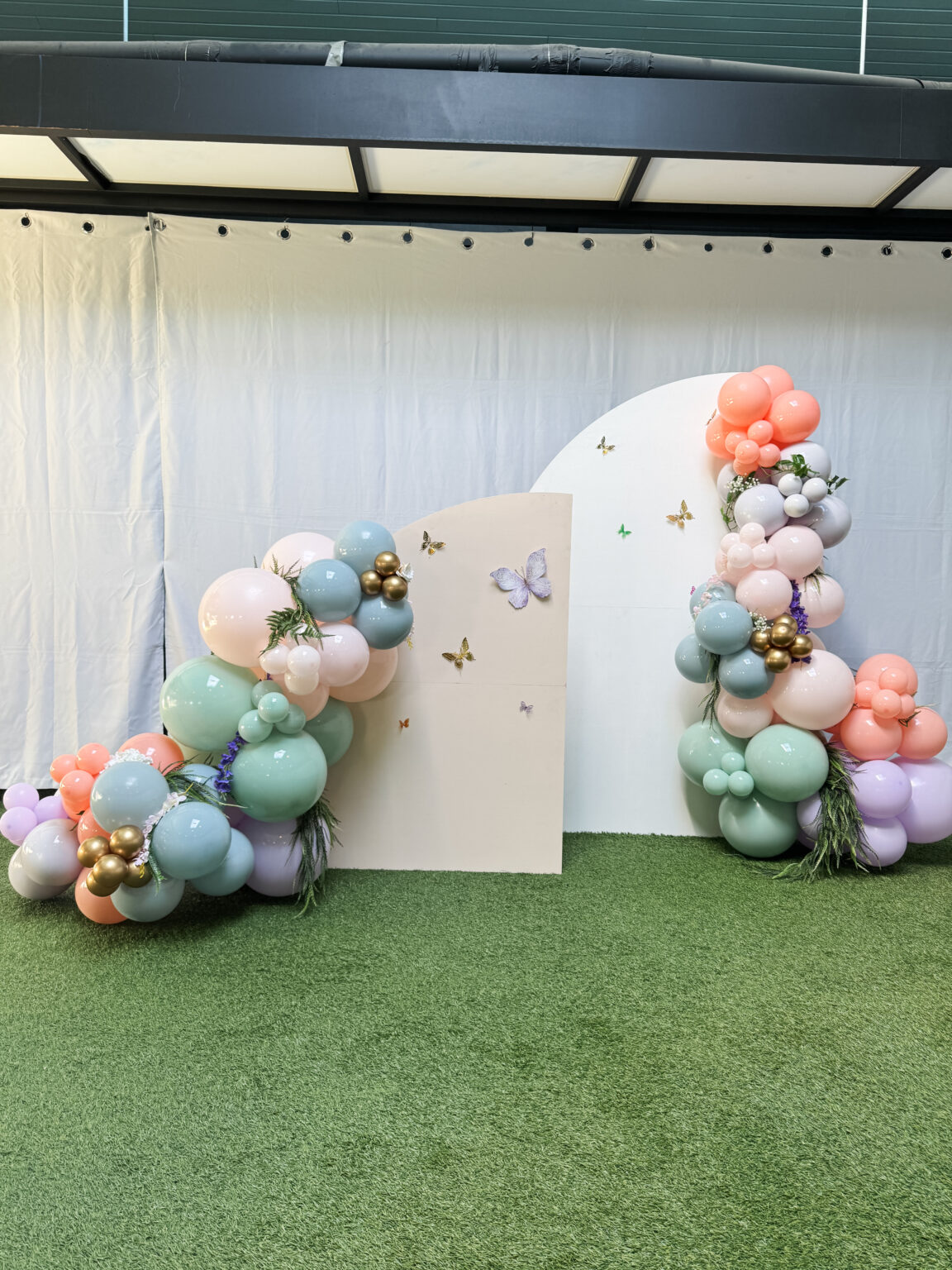 Pastel backdrop with butterfly accents, cream arch panels and sage, blush and lilac balloon garlands on the lawn