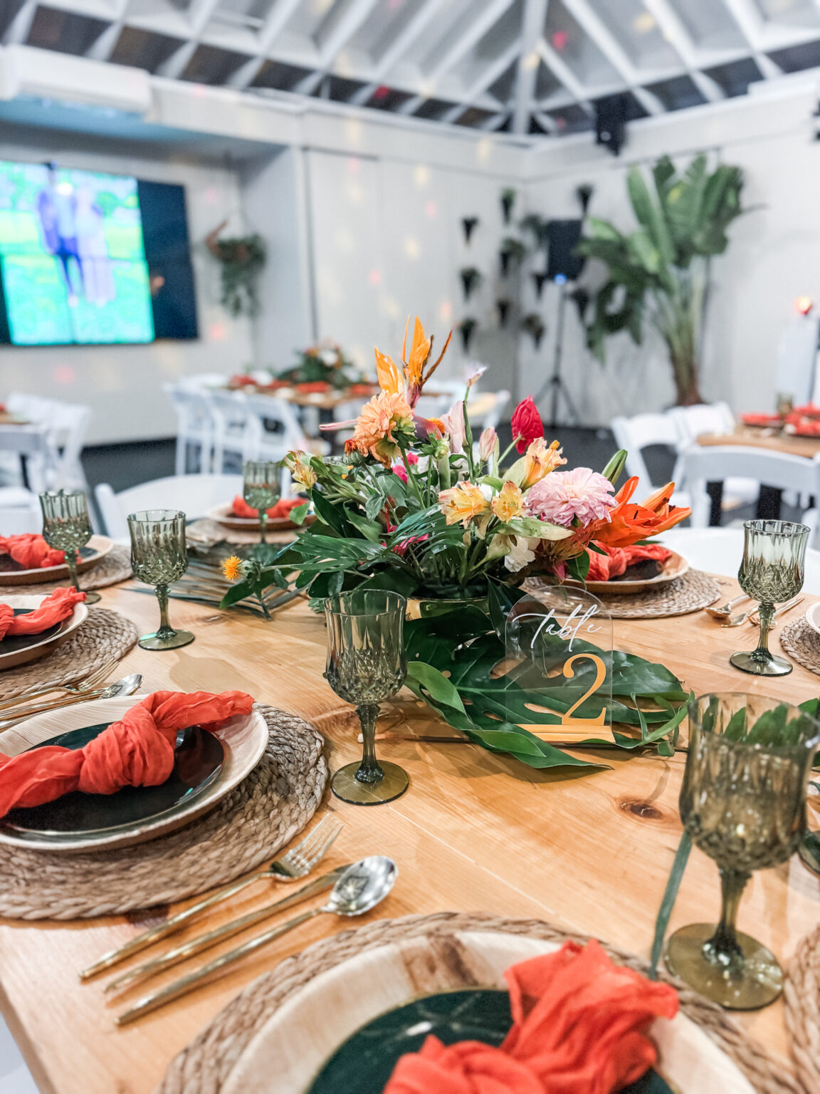 Tropical tablescape with a bird-of-paradise and dahlia centerpiece, monstera leaves, green crystal goblets and coral napkins on a wood farm table