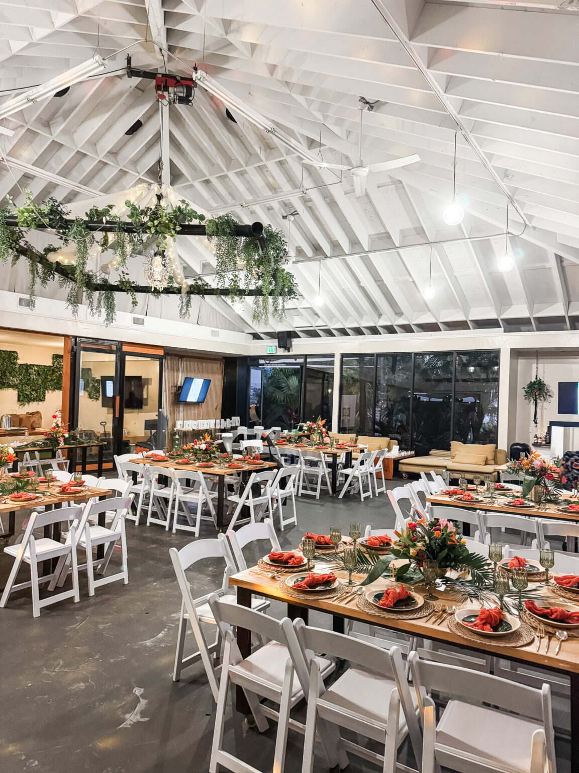 Indoor-outdoor venue set for a large celebration with long farm tables, white chairs, greenery garlands overhead and styled place settings