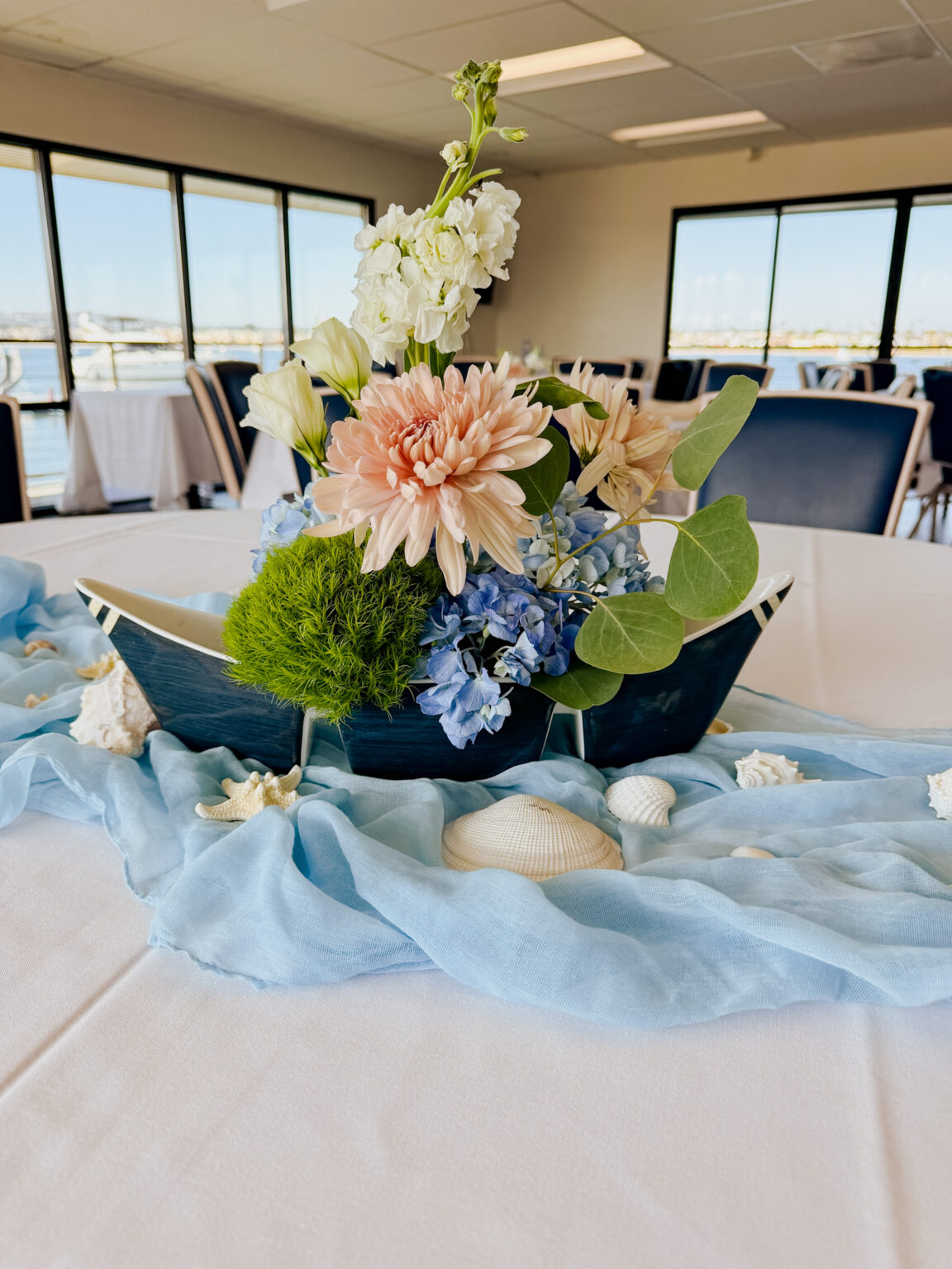 Floral centerpiece of peach dahlias, white blooms and blue hydrangea in a boat-shaped vessel on a dusty-blue runner