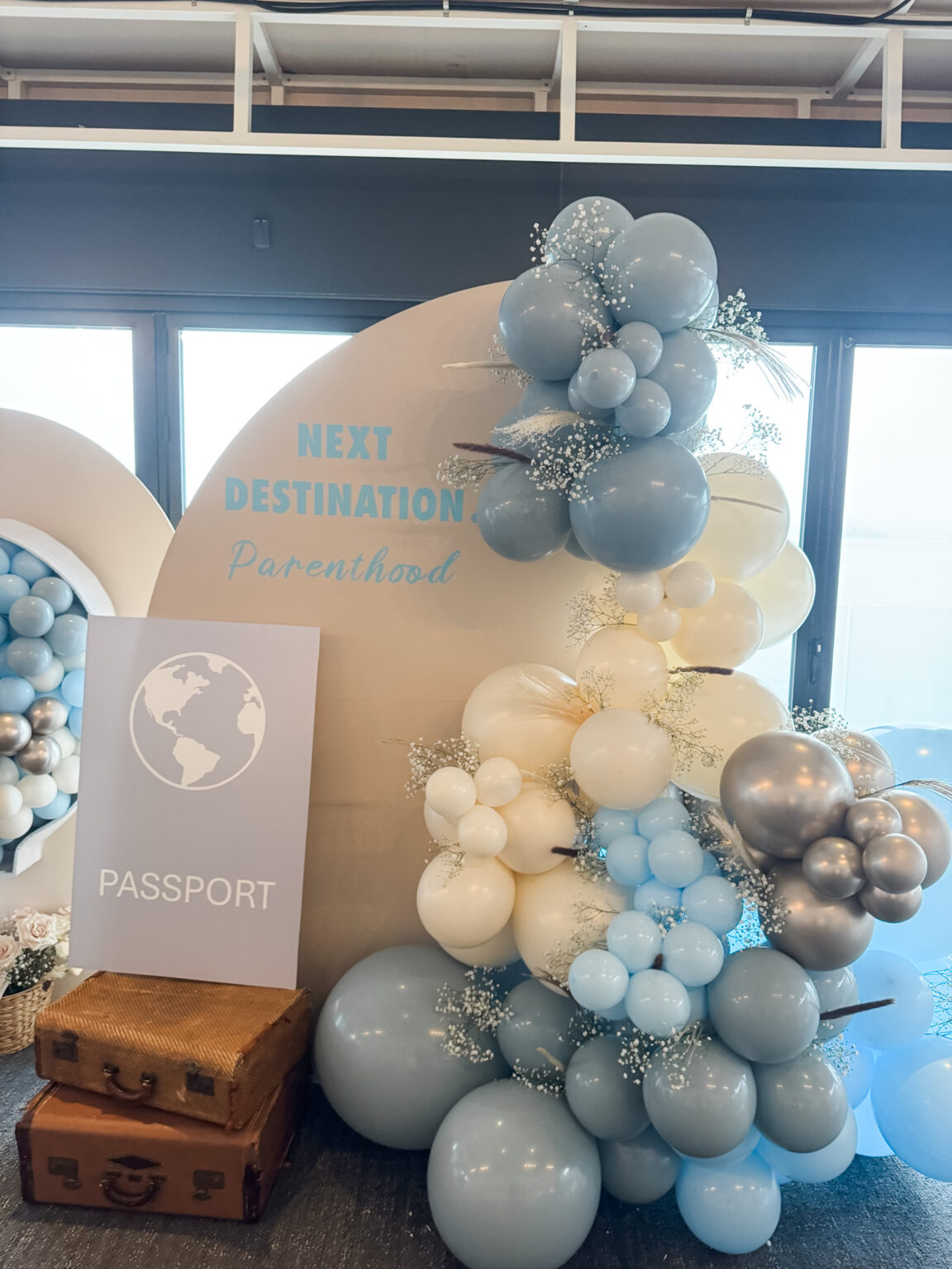 Travel-themed baby shower arch reading "Next Destination: Parenthood" with dusty-blue, cream and silver balloons, a passport sign and vintage suitcases