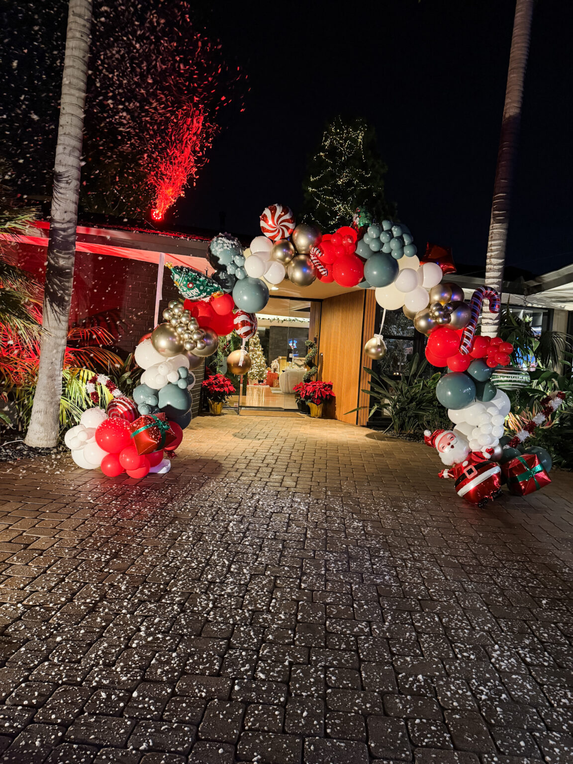 Holiday balloon arch in red, white, teal and silver with candy-cane and Santa accents framing a venue entrance at night