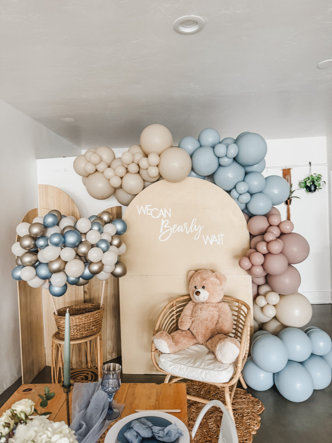Signature Gathering setup: tan, dusty-blue, mauve and silver balloon garland over two wood arch panels reading "We Can Bearly Wait," with a teddy bear in a rattan chair