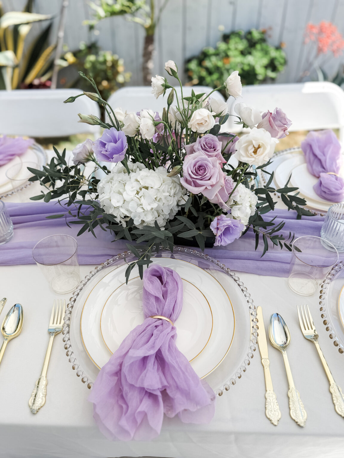 Layered tablescape with a lavender gauze runner and napkin, beaded glass chargers, gold-rimmed plates, gold flatware and a lavender rose and white hydrangea centerpiece