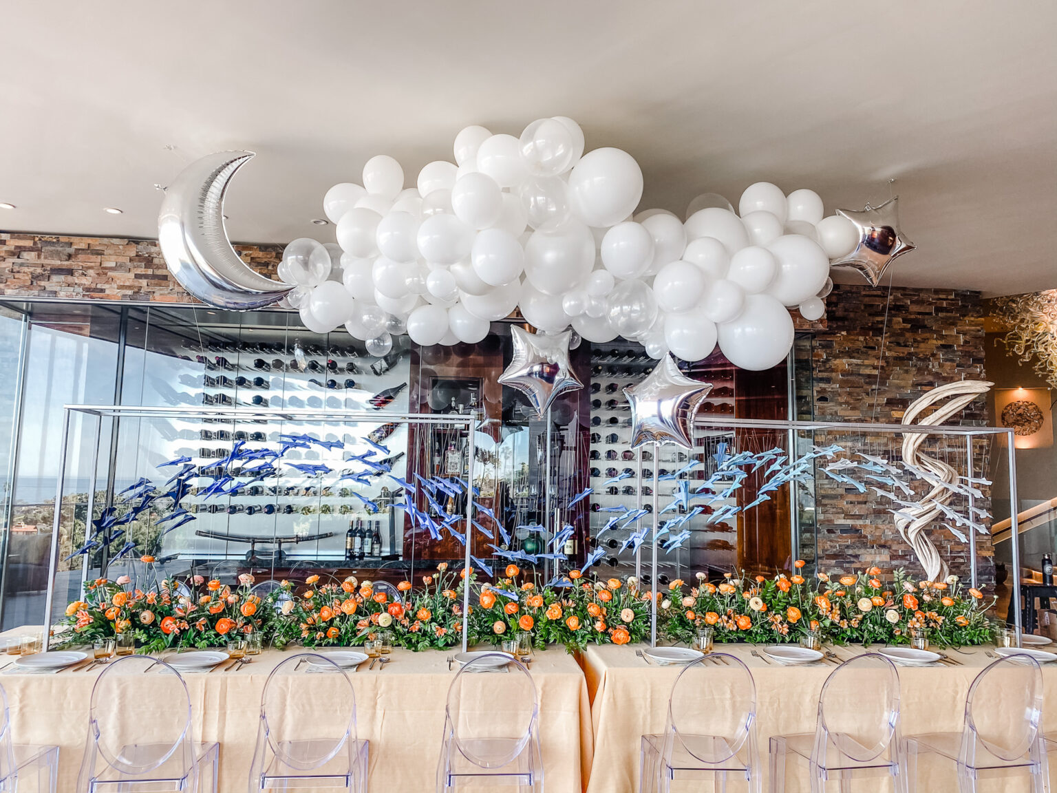 Long head table under a white balloon cloud with a silver moon and star balloons, clear frames filled with blue paper birds, an orange ranunculus garden runner and ghost chairs