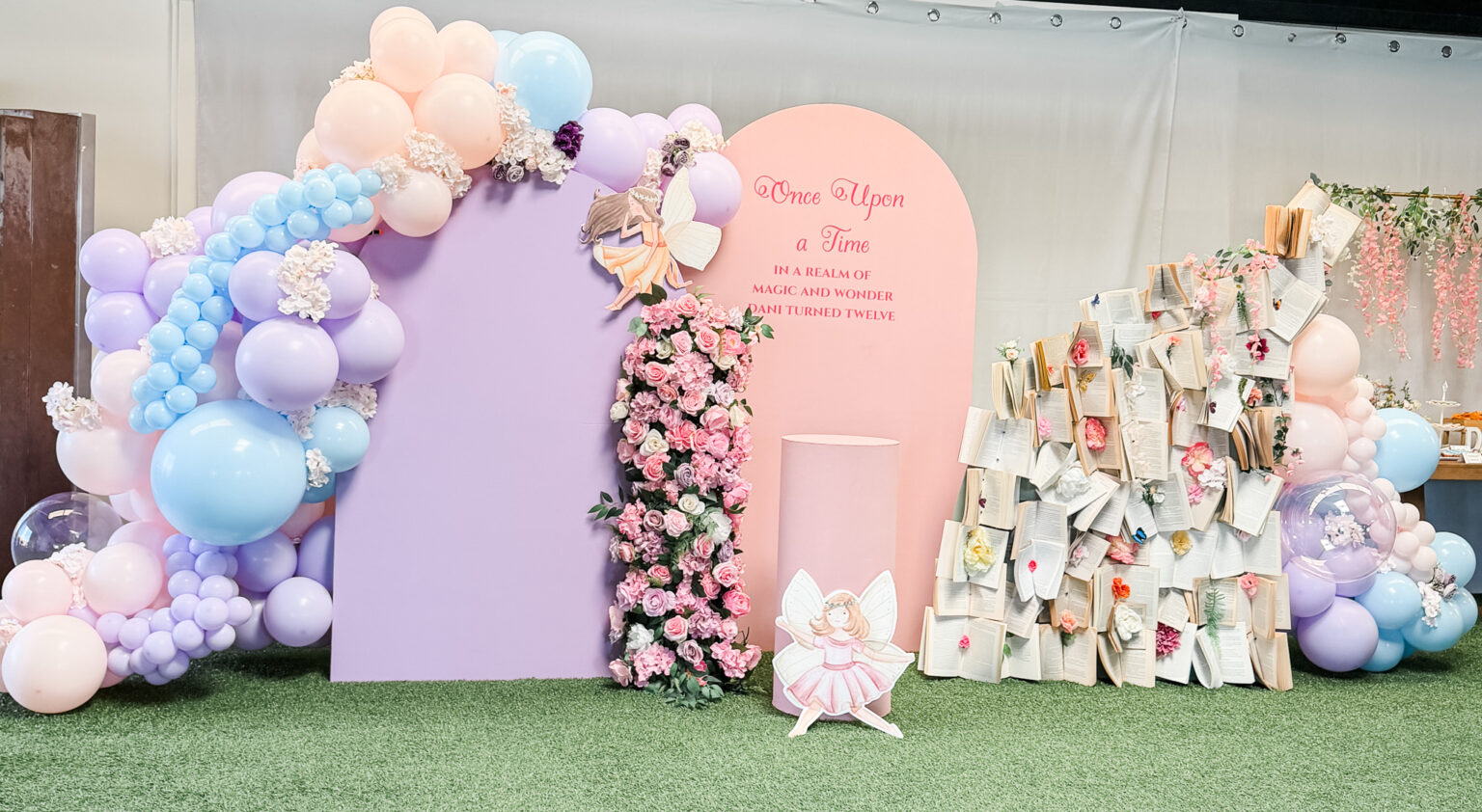 "Once Upon a Time" fairytale backdrop with lilac and pink arch panels, a pastel balloon garland, a pink floral column, a fairy cutout and a wall of open storybooks