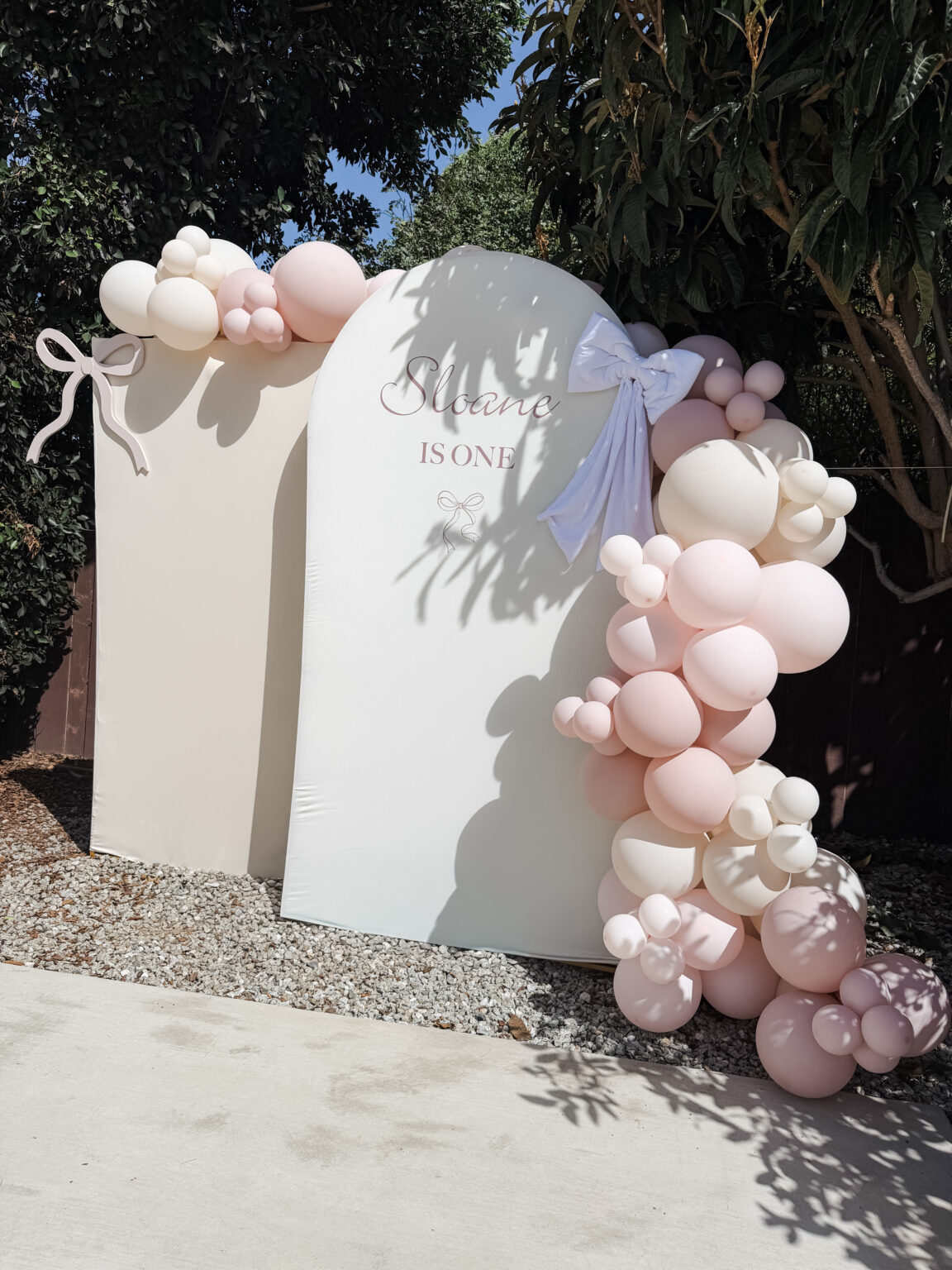 Two white arch panels reading "Sloane is One" with a blush satin bow and a blush and cream balloon garland, set outdoors under the trees