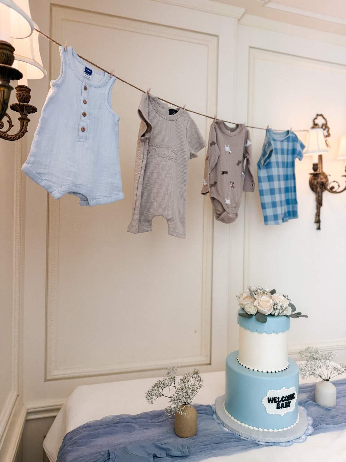 Baby clothes hung on a clothesline above a two-tier blue and white cake with white florals