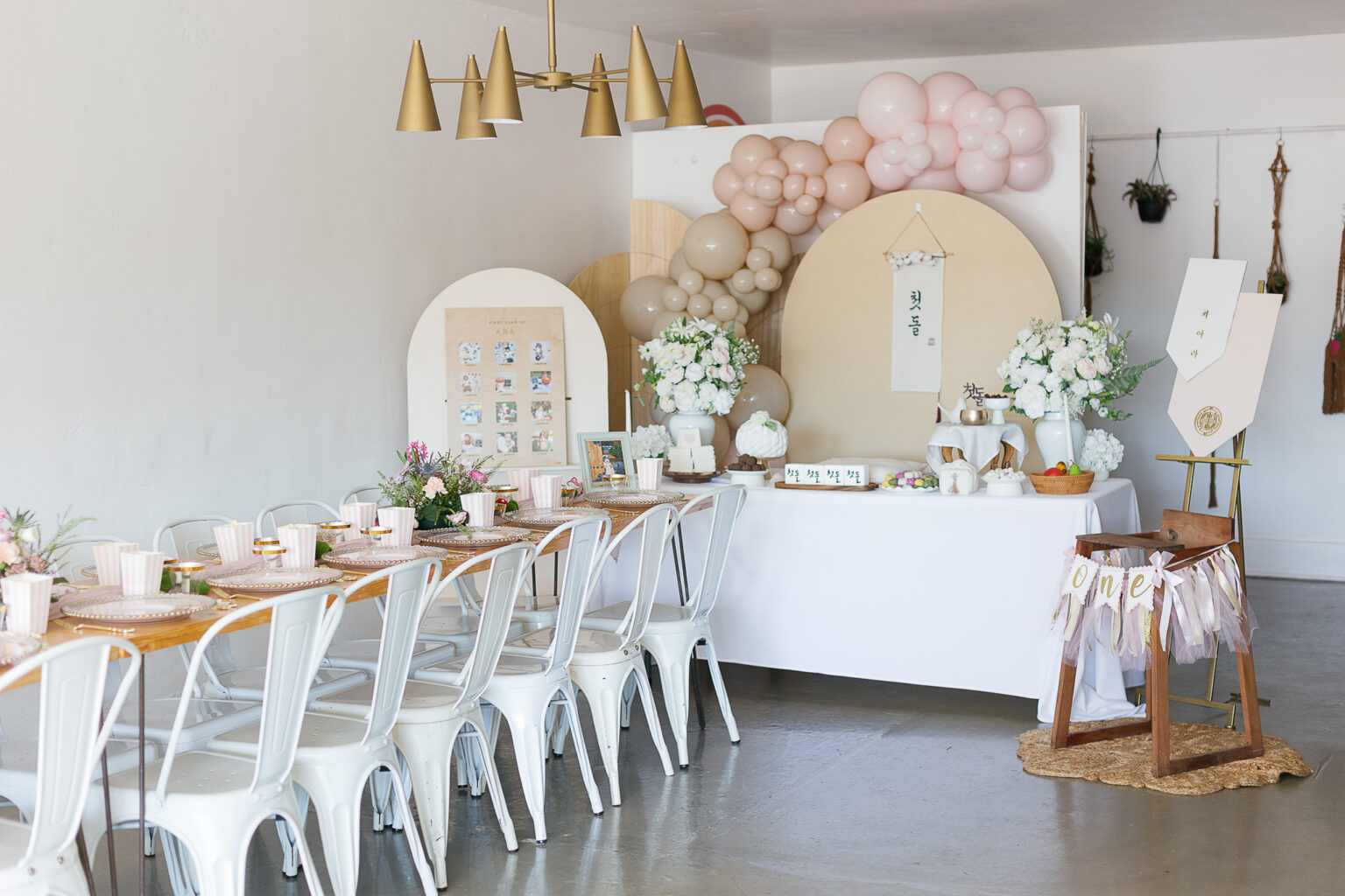 Korean first birthday (dol) celebration with a long table of white chairs, a neutral arch backdrop with blush and taupe balloons, white florals on the dessert table and a ribboned high chair