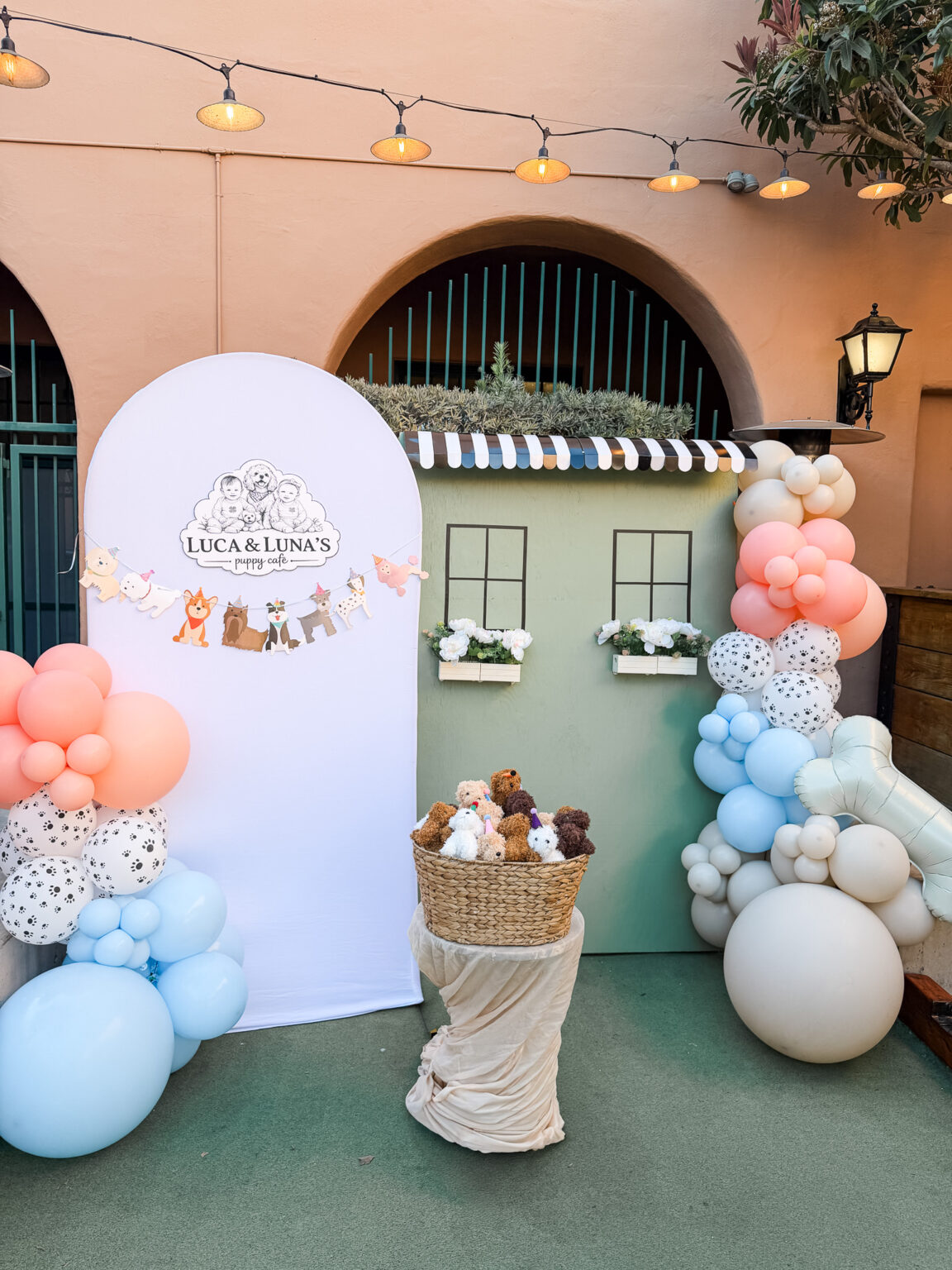 Outdoor baby shower installation with a custom arch panel, a dimensional storefront facade and blush, blue and cow-print balloon garlands