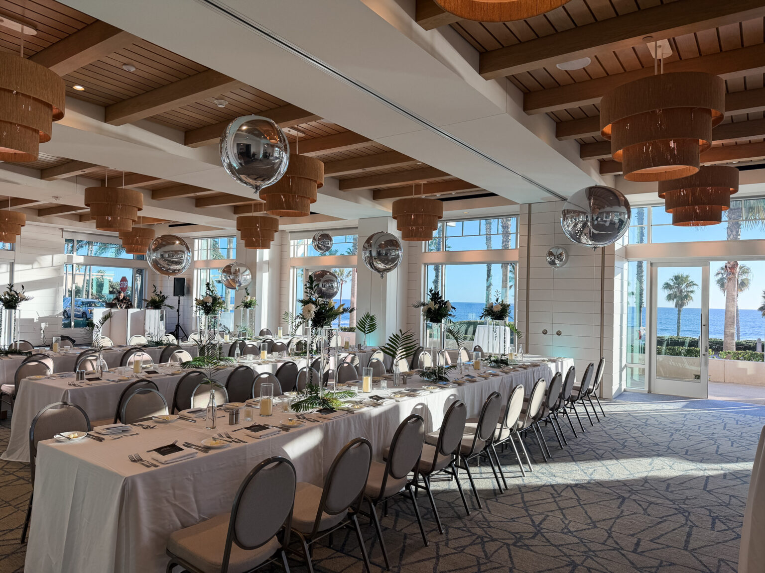 Bright waterfront ballroom set for a large celebration with long draped banquet tables, place settings and greenery