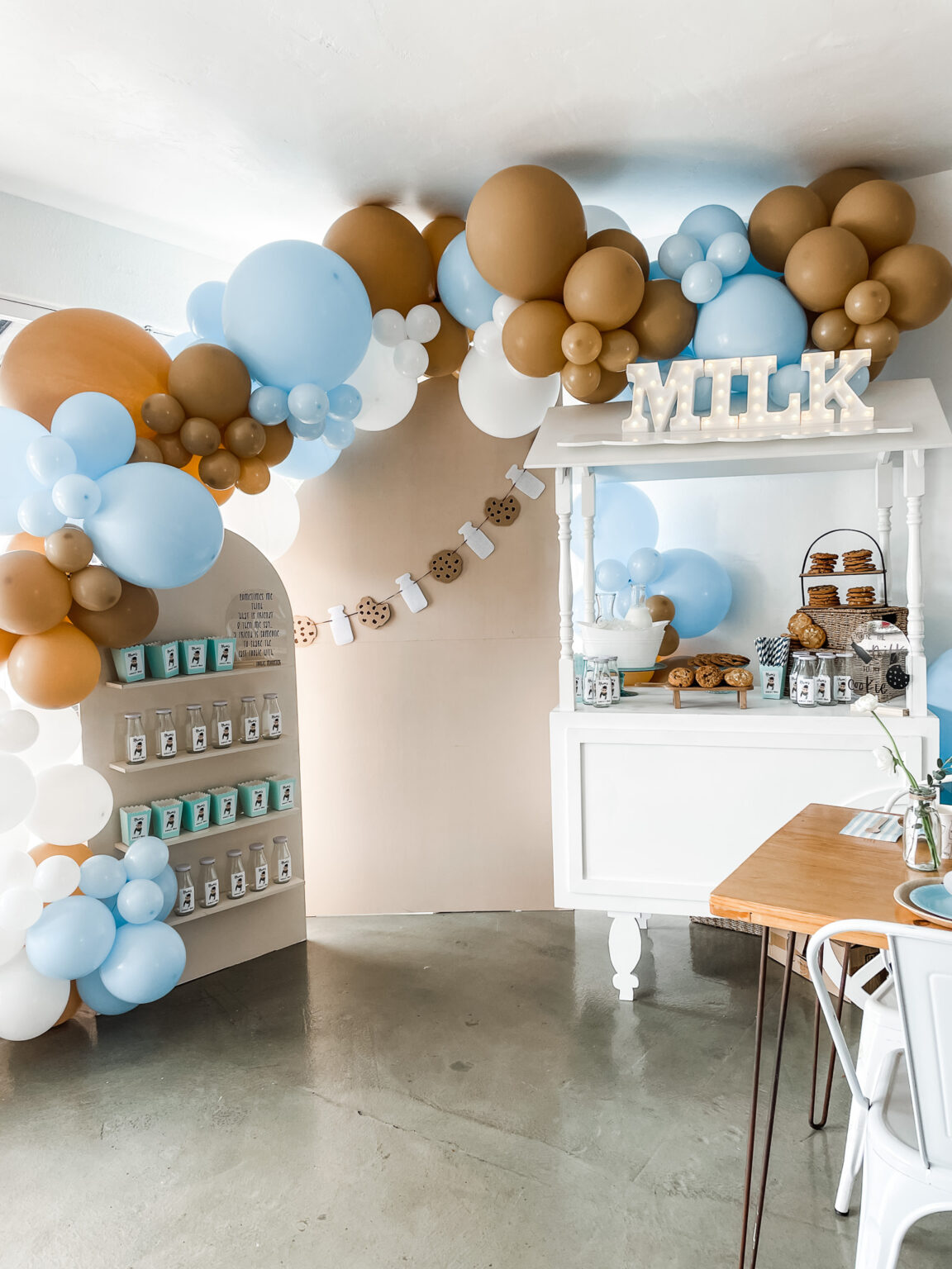 Milk-and-cookies first birthday installation with an oversized organic balloon garland in caramel, sky blue and white over a cookie banner and milk bottle display