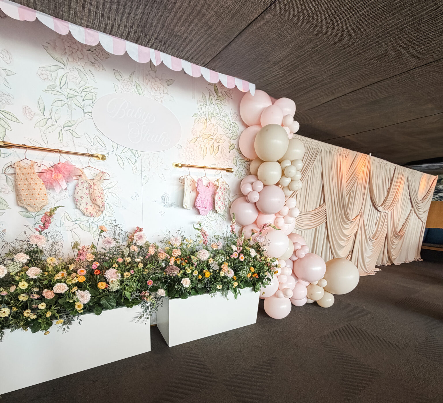 Long event backdrop with a soft floral mural, blush and cream balloon garland and planter boxes of fresh flowers