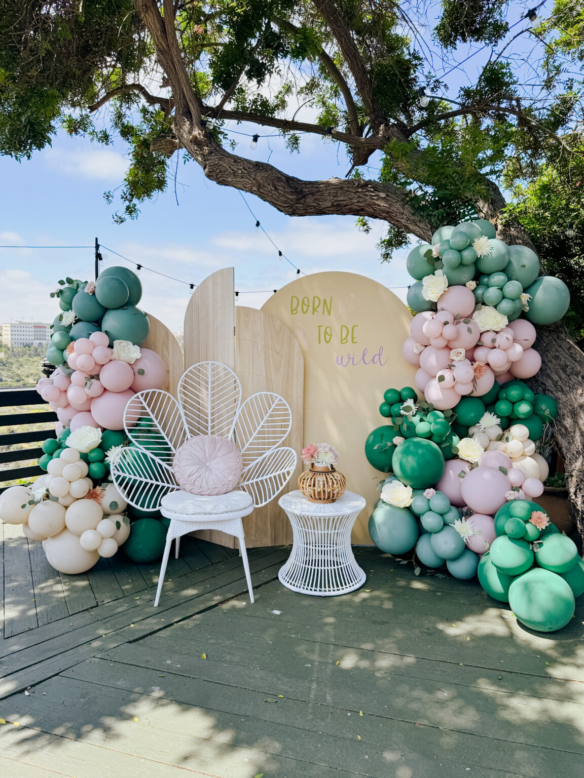 Outdoor celebration with pastel pink, lilac and teal balloon garlands around two arch panels and a white peacock chair under a tree