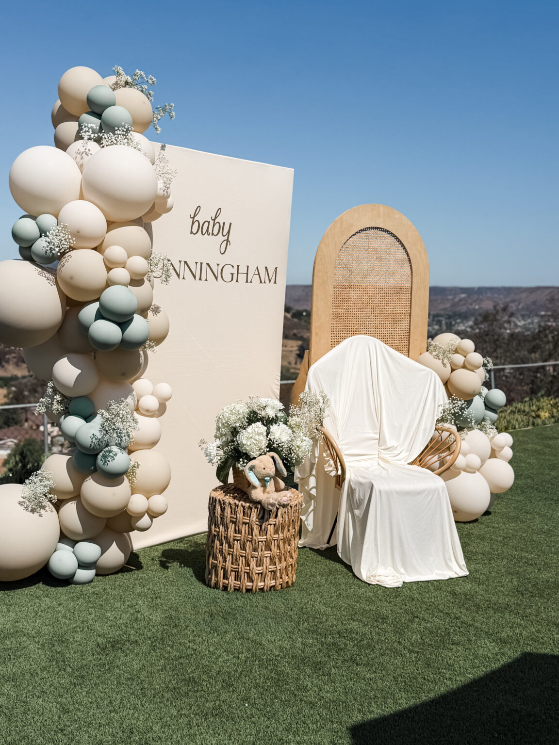 Baby shower on a hilltop lawn: cream and sage balloon garland, a "baby Cunningham" welcome panel, rattan arch backdrop and draped rattan chair with canyon views behind