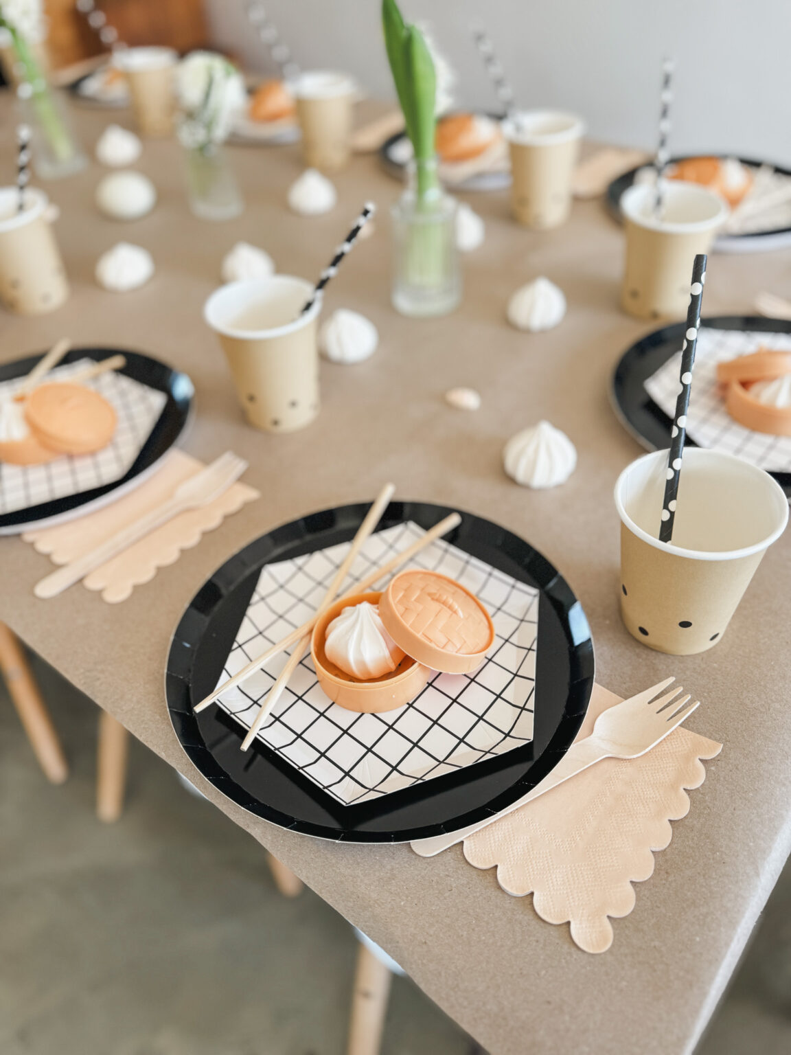 Neutral kids' table setting with black plates, grid napkins, kraft cups with striped straws, scalloped wooden cutlery and meringue treats
