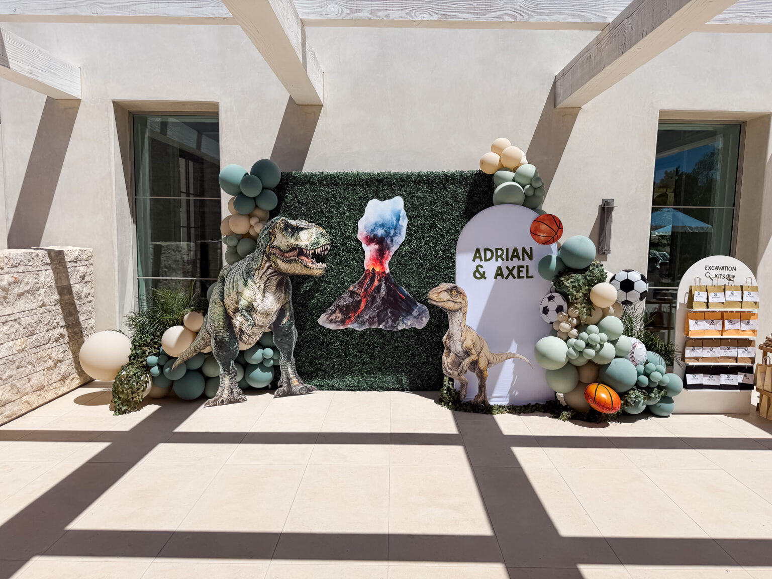 Dinosaur birthday installation with a life-size T-rex, greenery wall, custom name arch panels and sage, cream and rust balloon garlands