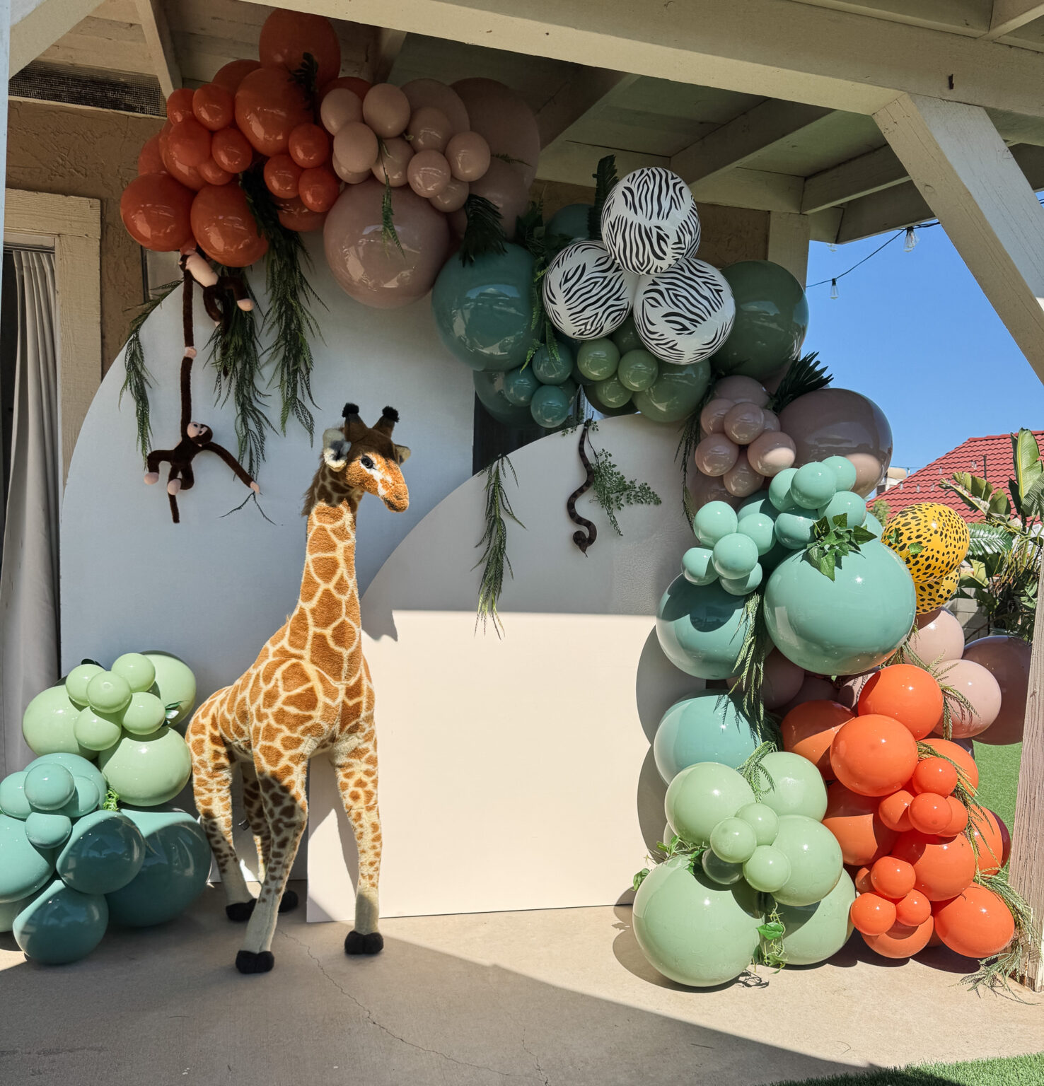 Safari-themed setup with a giraffe figure, sage, terracotta and patterned balloons and a rounded backdrop under a covered patio