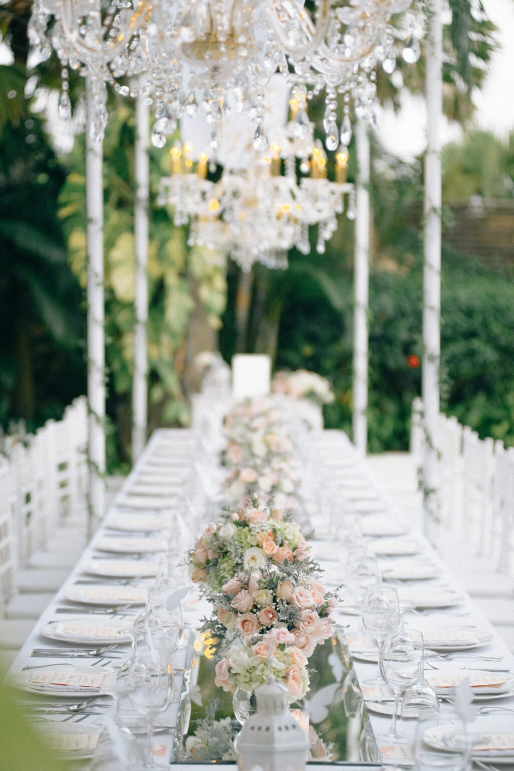 Long garden wedding reception table under crystal chandeliers with blush and white florals and candlelight