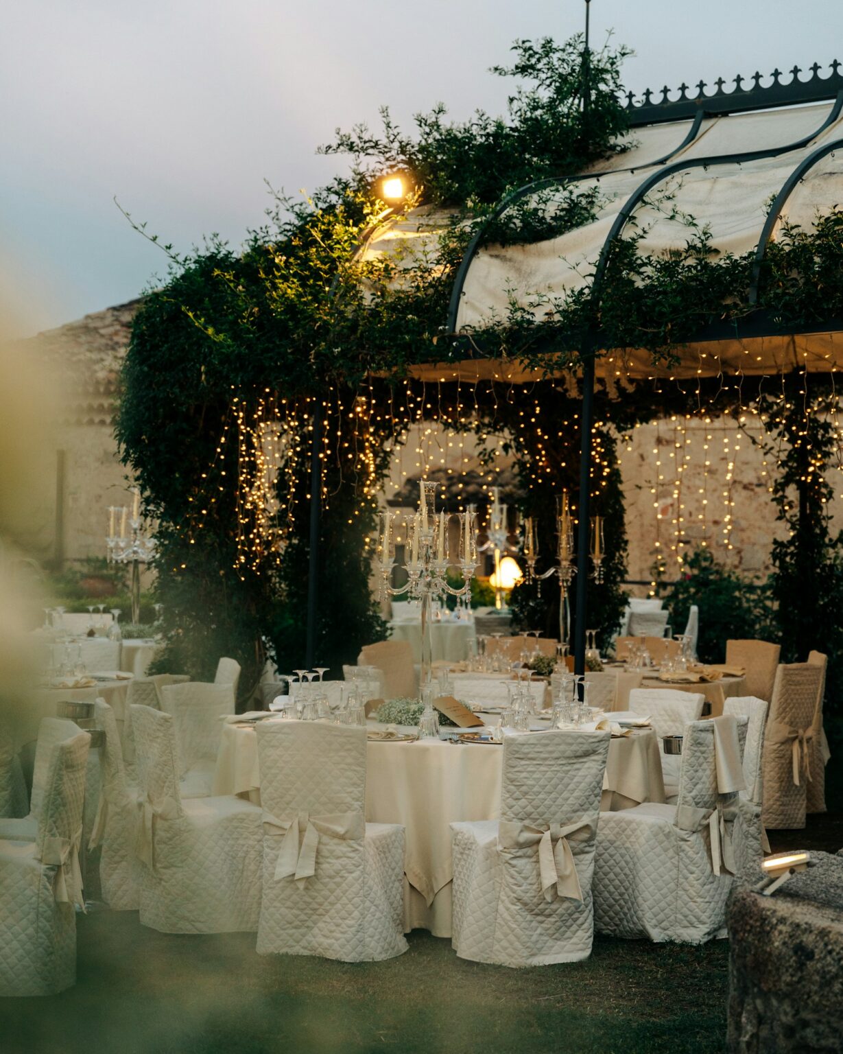 Garden wedding reception at dusk with white-draped tables and chairs under string lights and greenery